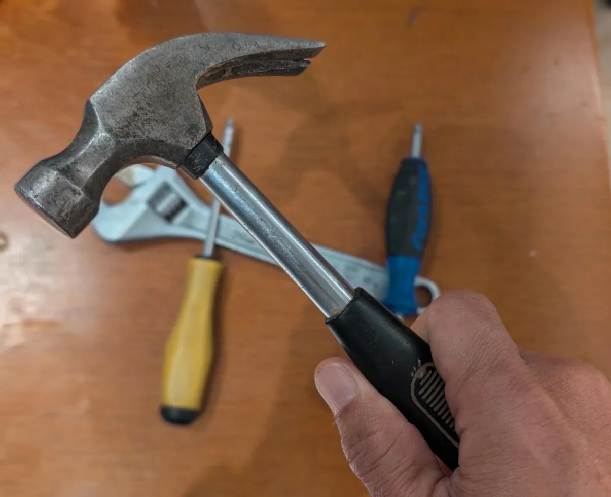 A hand holding a hammer above tools on a wooden workbench.